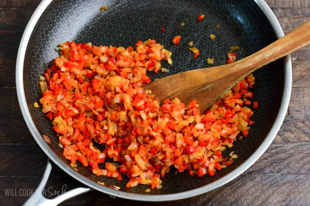 Finely diced red bell peppers and onions sautéing in a skillet with a wooden spoon, cooking down for turkey meatloaf.