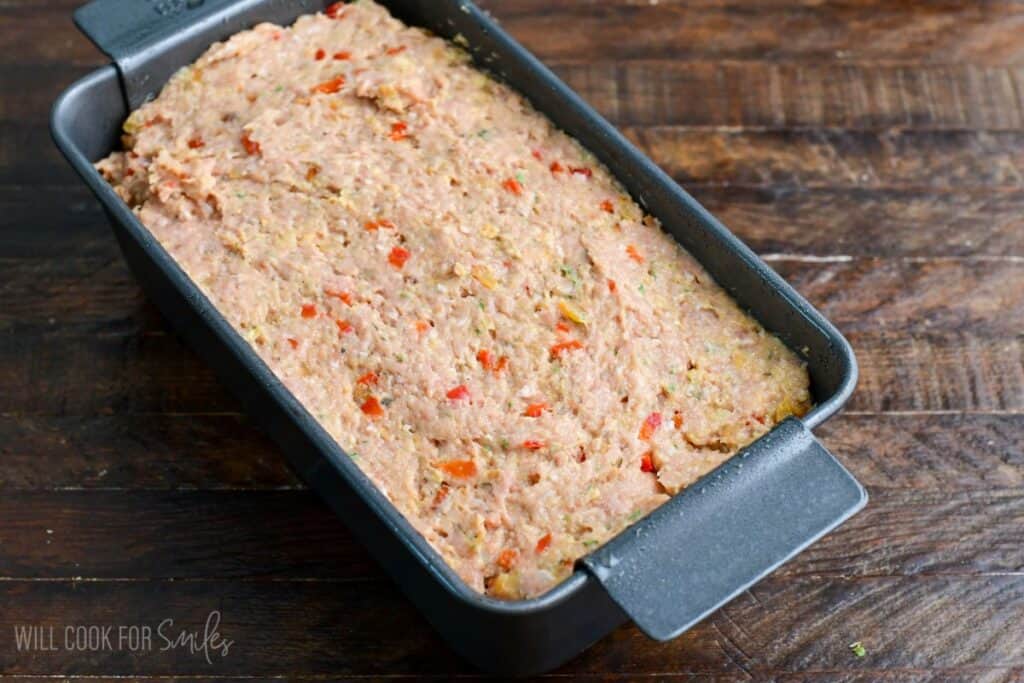 Uncooked turkey meatloaf mixture pressed evenly into a loaf pan, ready to bake, with visible diced red peppers and herbs throughout.
