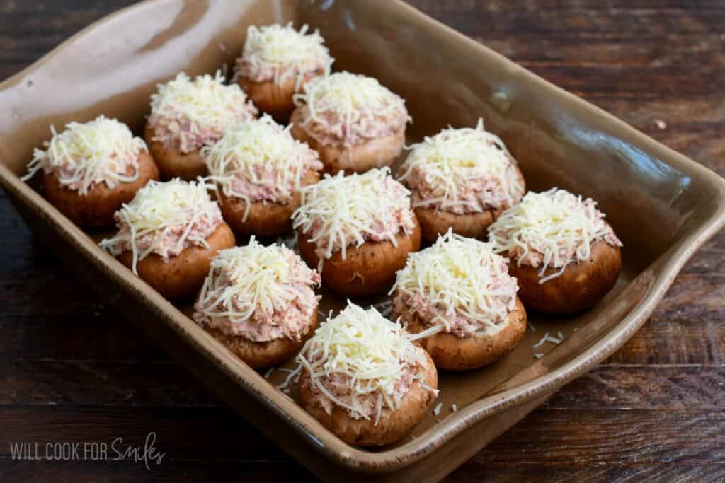 Baking dish filled with mushroom caps stuffed with creamy Reuben filling and topped with shredded Swiss cheese before baking.