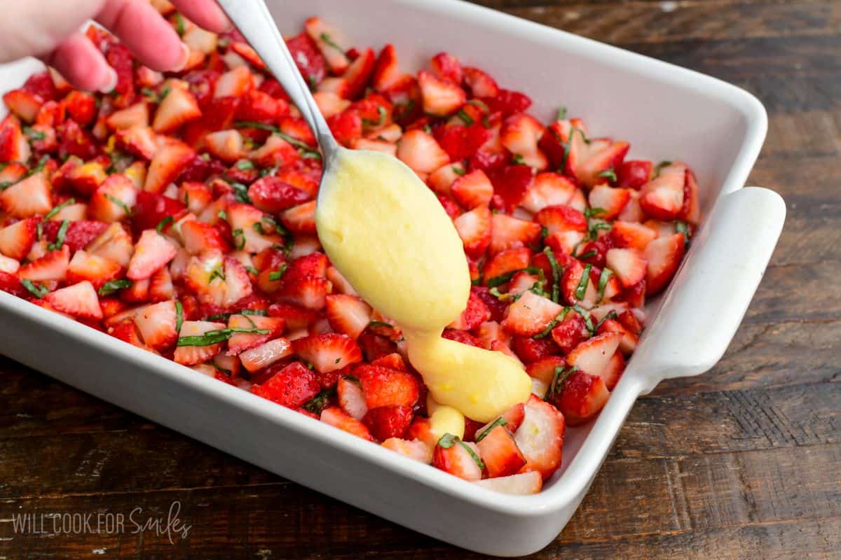 Spoonful of cake batter being dropped over chopped strawberries and basil in a baking dish for cobbler.