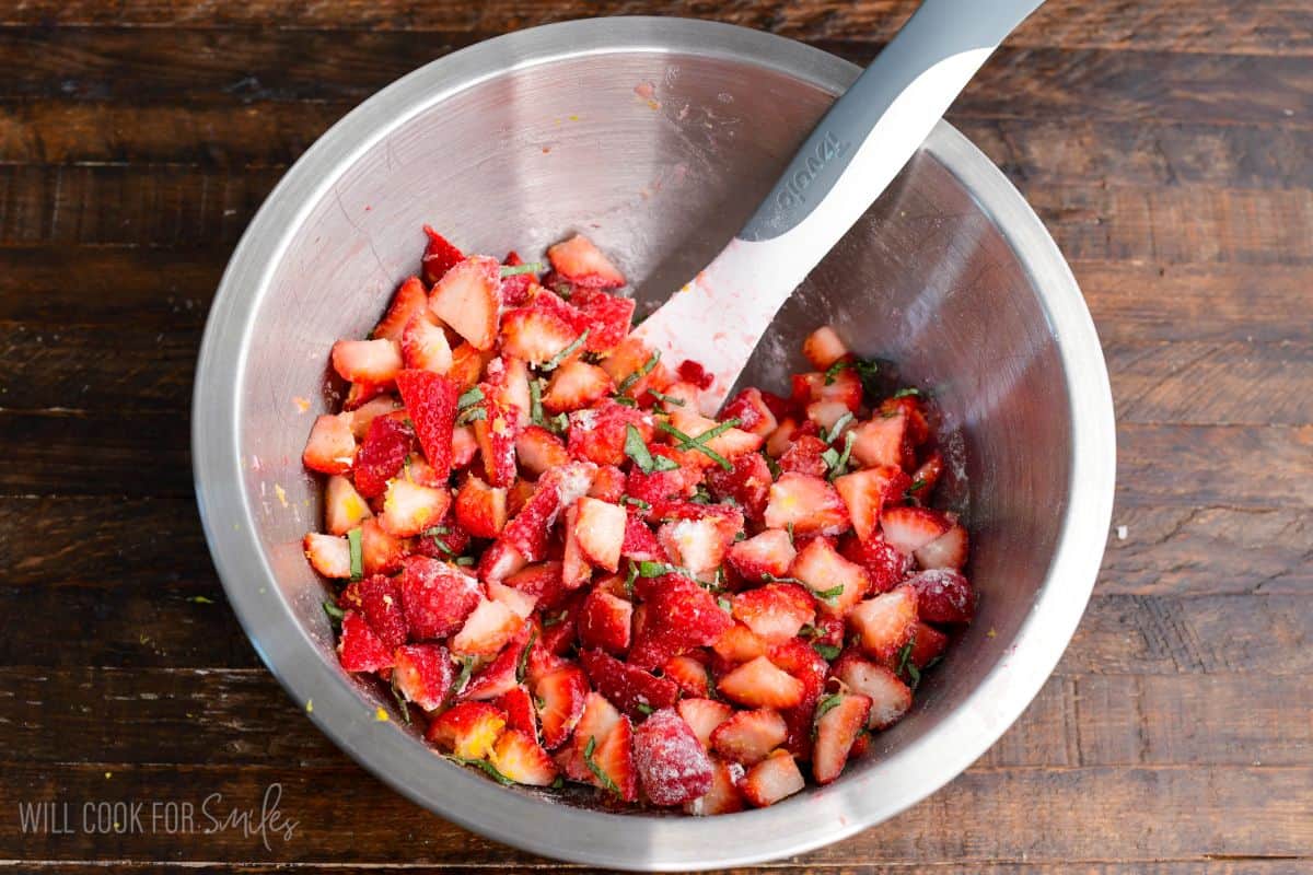 Mixing bowl with chopped strawberries tossed with sugar and fresh basil, being stirred with a spatula.