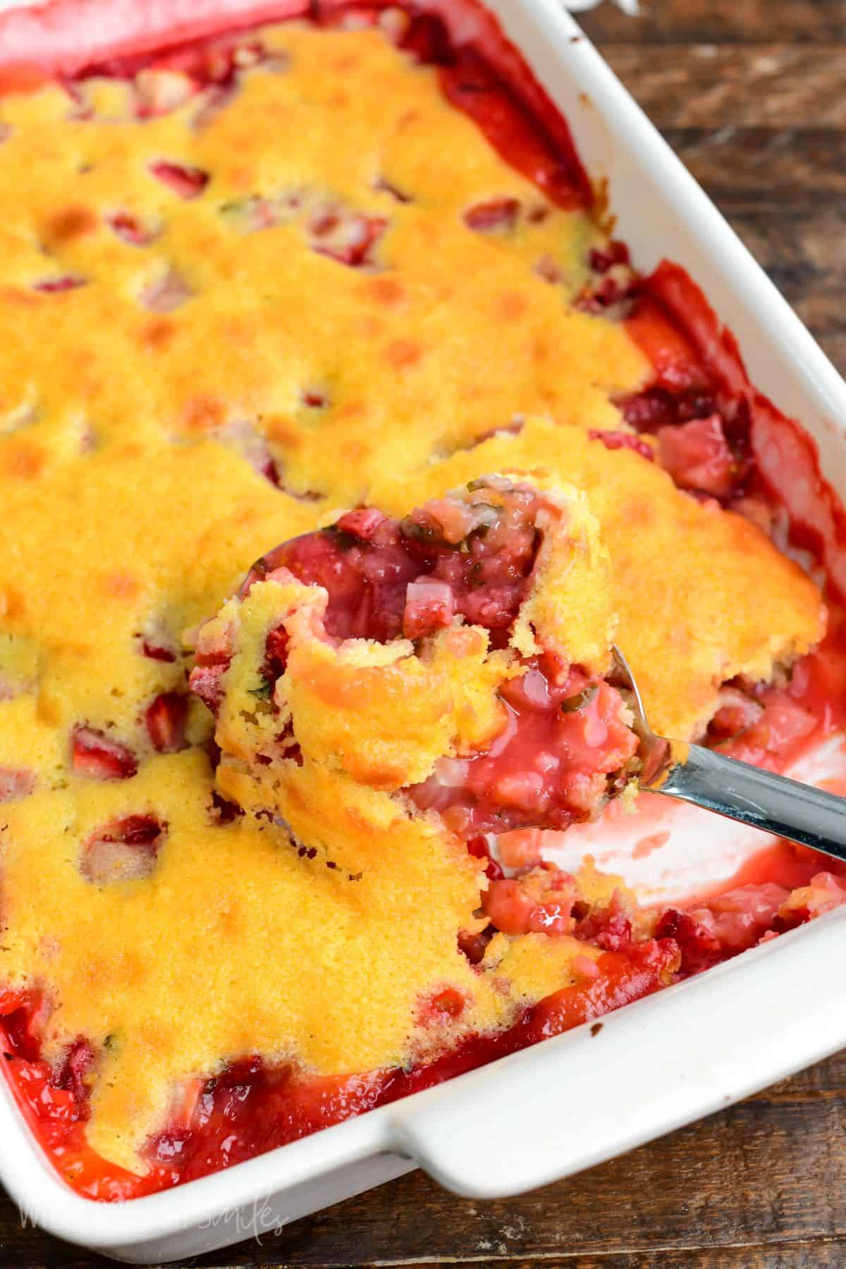 Spoon scooping a serving of strawberry basil cobbler from a white baking dish, showing the juicy strawberry filling and soft golden cake topping.