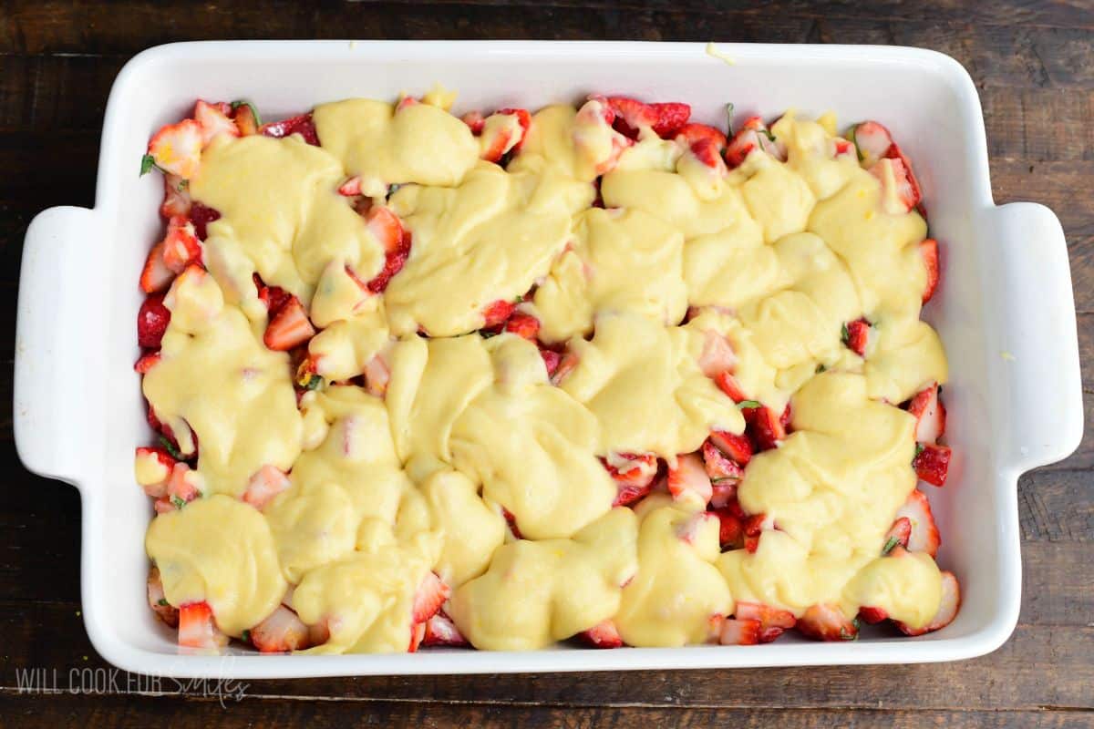 Baking dish with strawberries and basil topped with dollops of cake batter before baking into cobbler.