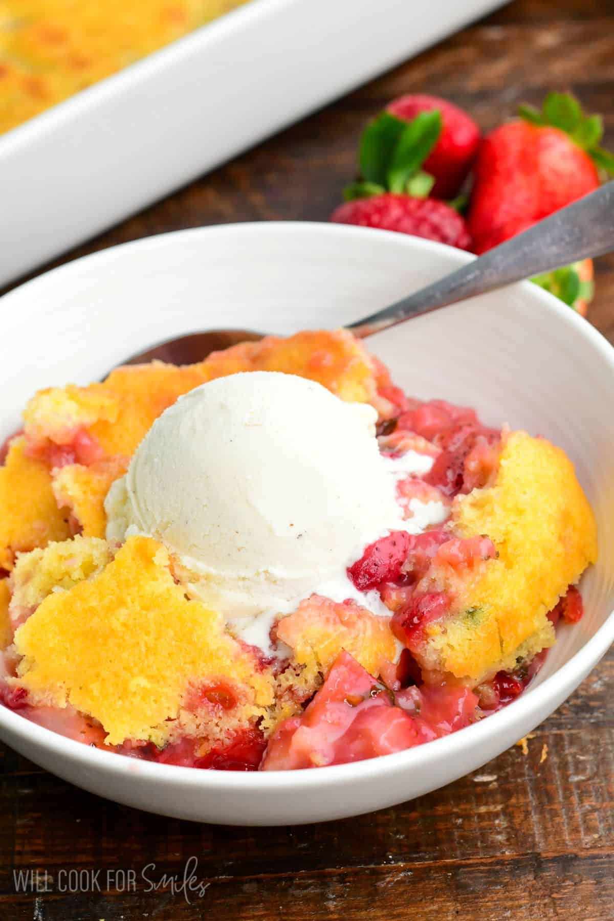Close-up of strawberry basil cobbler in a bowl topped with a scoop of vanilla ice cream, with a spoon and fresh strawberries in the background.