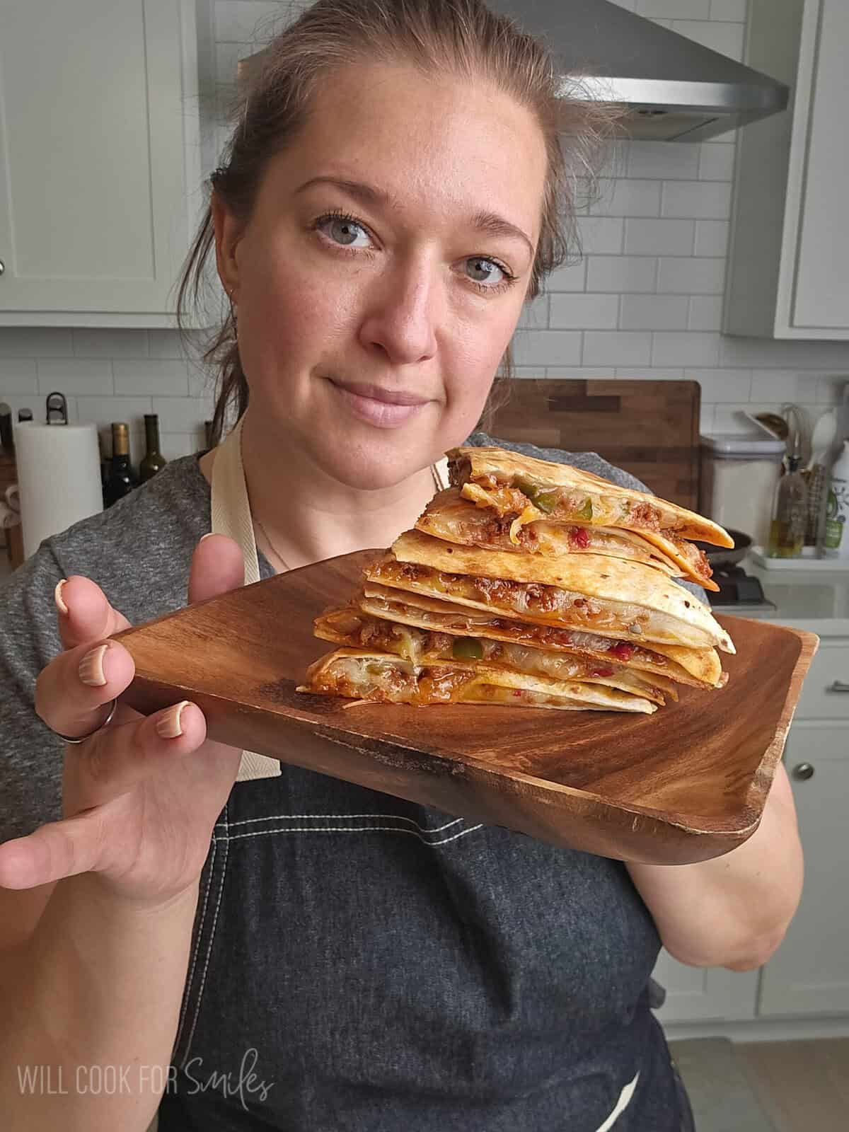 Person holding a wooden board with stacked cheesy quesadilla slices filled with meat and vegetables in a kitchen.
