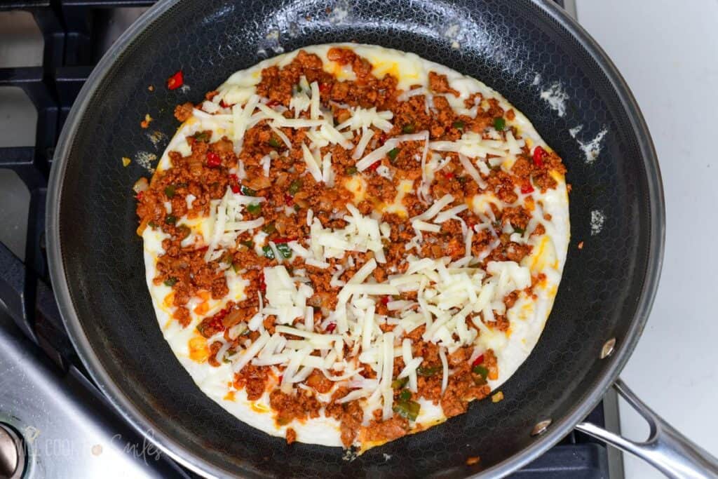 Chorizo and shredded cheese layered on a tortilla cooking in a skillet on the stovetop.