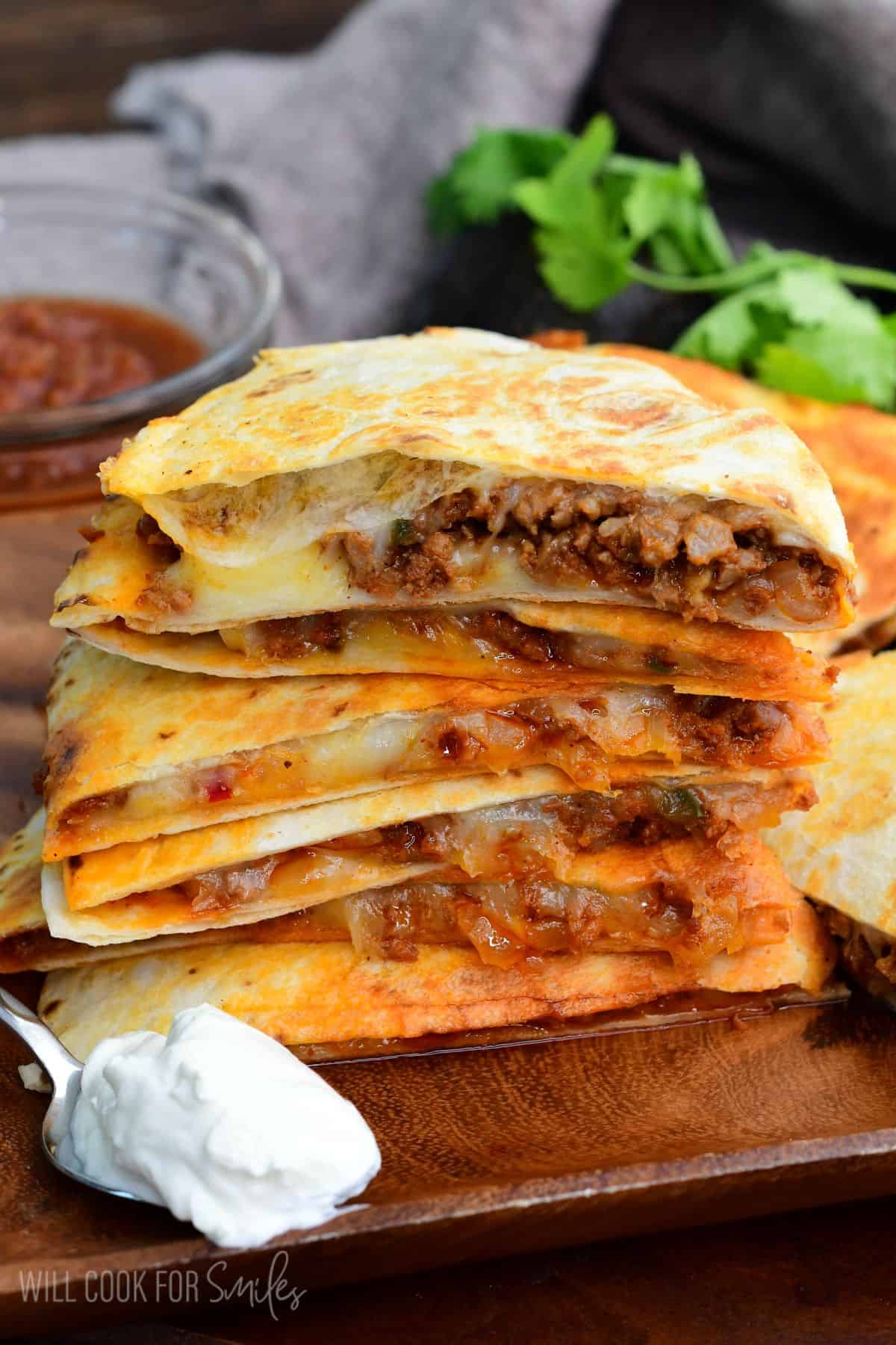 Stacked chorizo quesadilla slices with melted cheese and seasoned meat filling, served with sour cream on a wooden board.