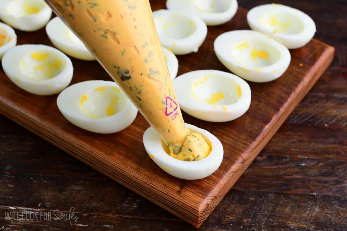 Deviled eggs being filled with a creamy yolk mixture using a piping bag on a wooden board.