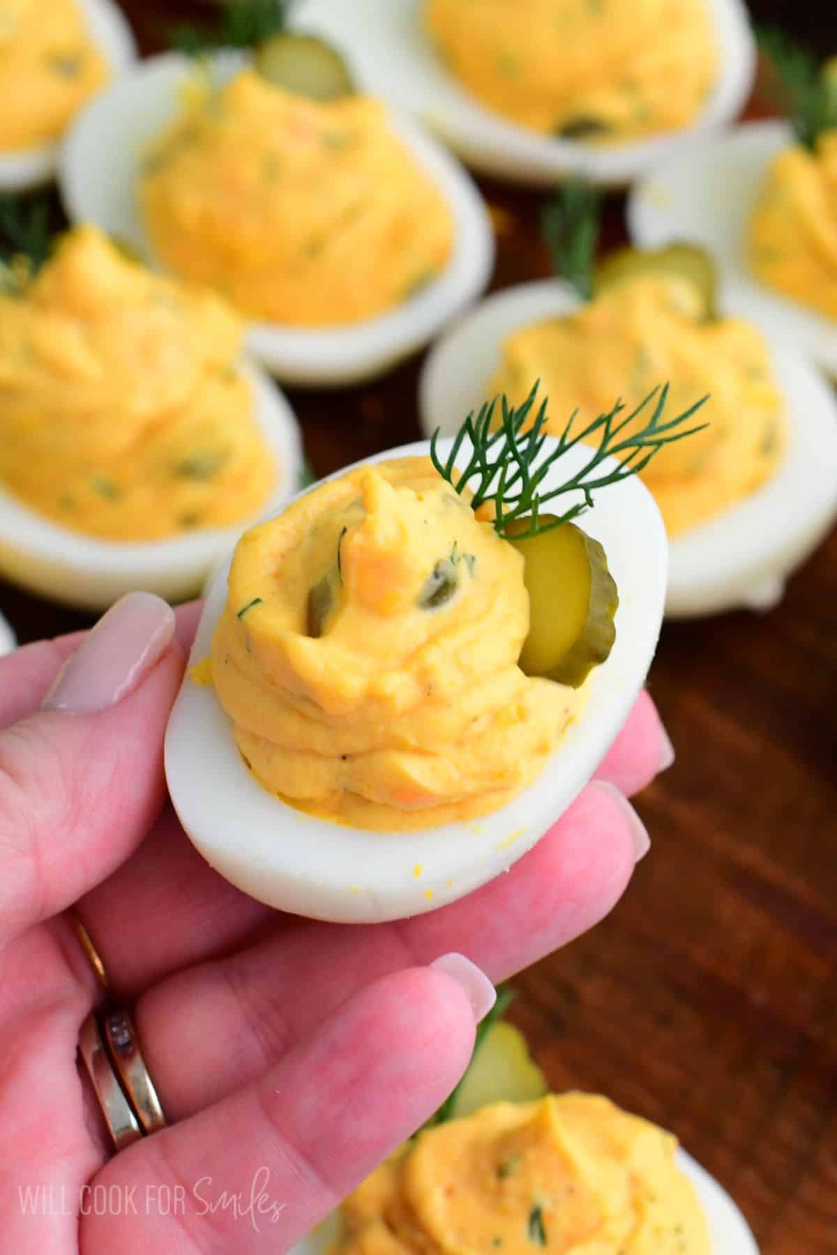 Hand holding a dill pickle deviled egg topped with creamy filling, a pickle slice, and fresh dill garnish.