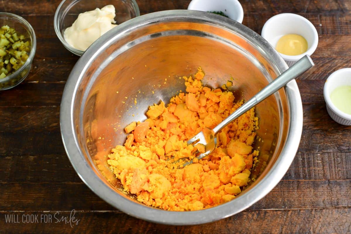 Egg yolks being mashed with a fork in a metal mixing bowl, with ingredients like mayonnaise, mustard, and chopped pickles nearby.
