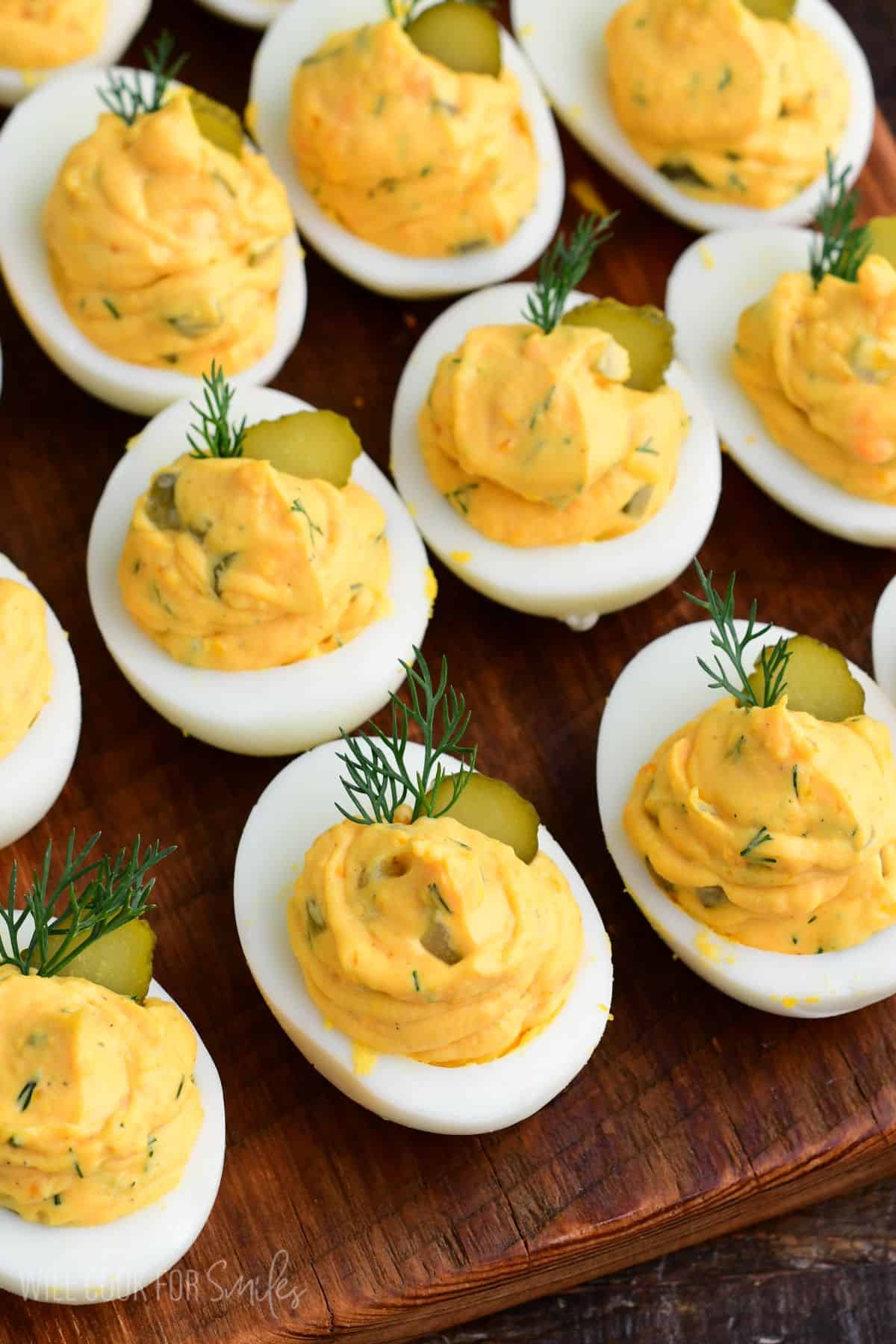 Close-up of dill pickle deviled eggs with creamy filling, topped with pickle slices and fresh dill on a wooden board.