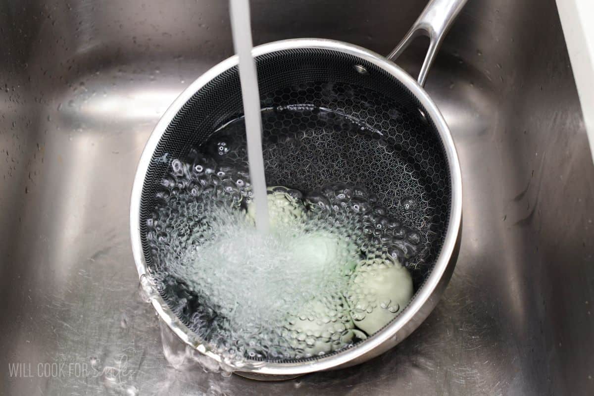 Hard-boiled eggs cooling in a saucepan under running cold water in a kitchen sink.