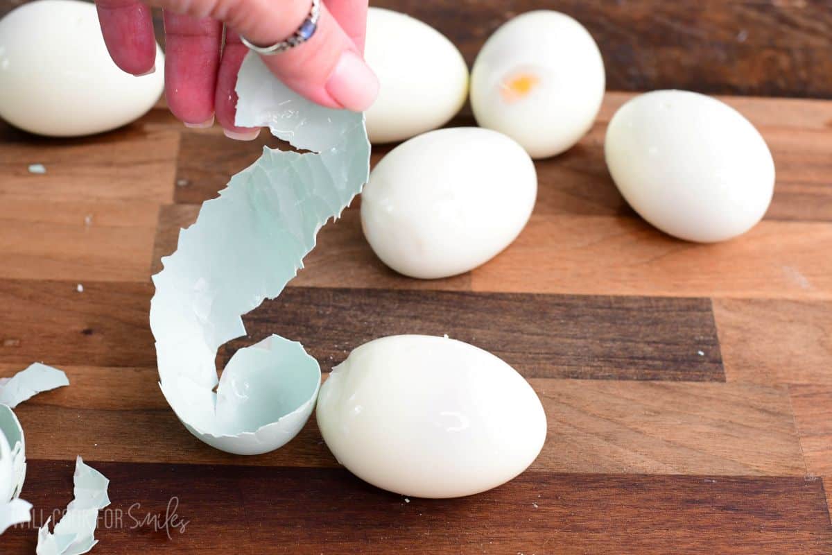 Hand removing a large piece of shell from a hard-boiled egg on a wooden cutting board, with several peeled eggs in the background.