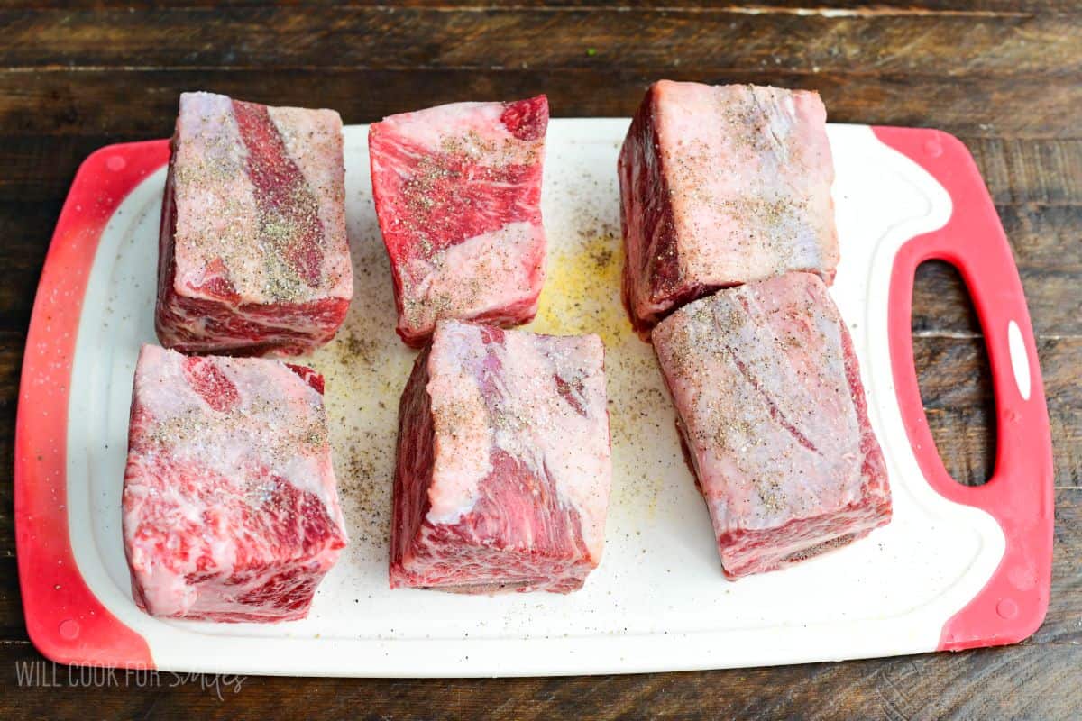six short ribs seasoned with salt and pepper on a white cutting board.