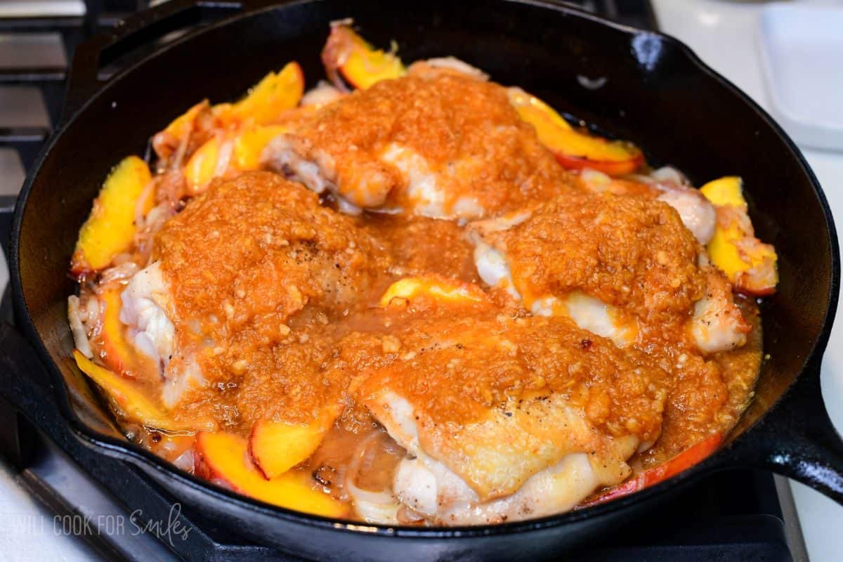 Chicken thighs topped with peach sauce and fresh peaches in a cast-iron skillet, ready to bake.