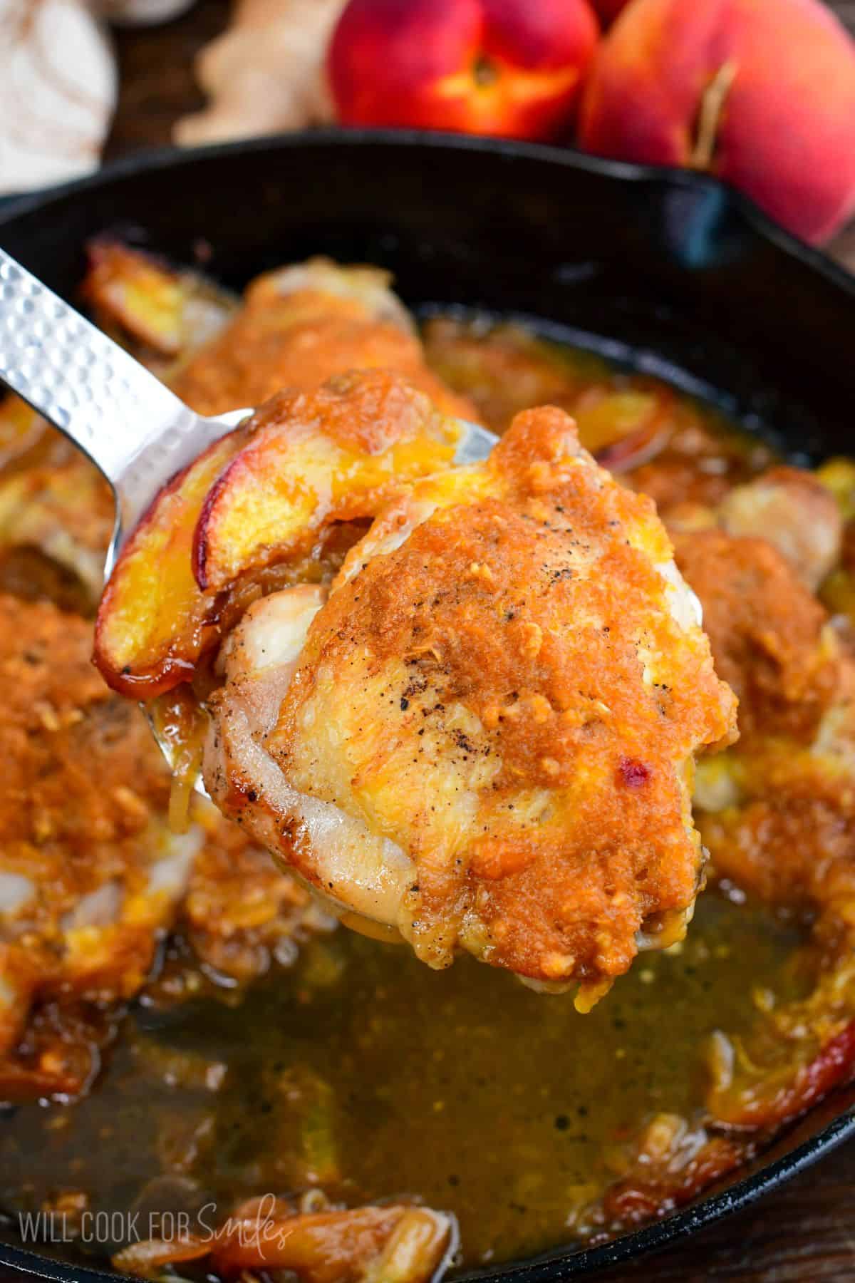 Spoon lifting a baked peach chicken thigh from a skillet with juicy peaches and savory peach onion sauce.