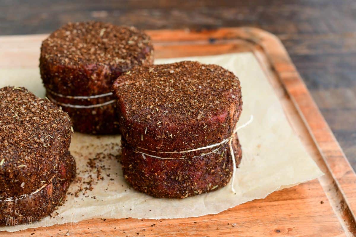 Coffee-rubbed filet mignon steaks tied with kitchen twine and coated in seasoning, resting on parchment paper atop a wooden cutting board.