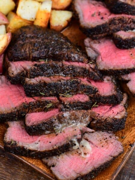 Sliced coffee-rubbed filet mignon arranged on a wooden serving platter, featuring a tender medium-rare center and a rich, dark crust. Melted herb butter and savory juices coat the steak slices, while crispy roasted potatoes add a hearty side dish in the background.