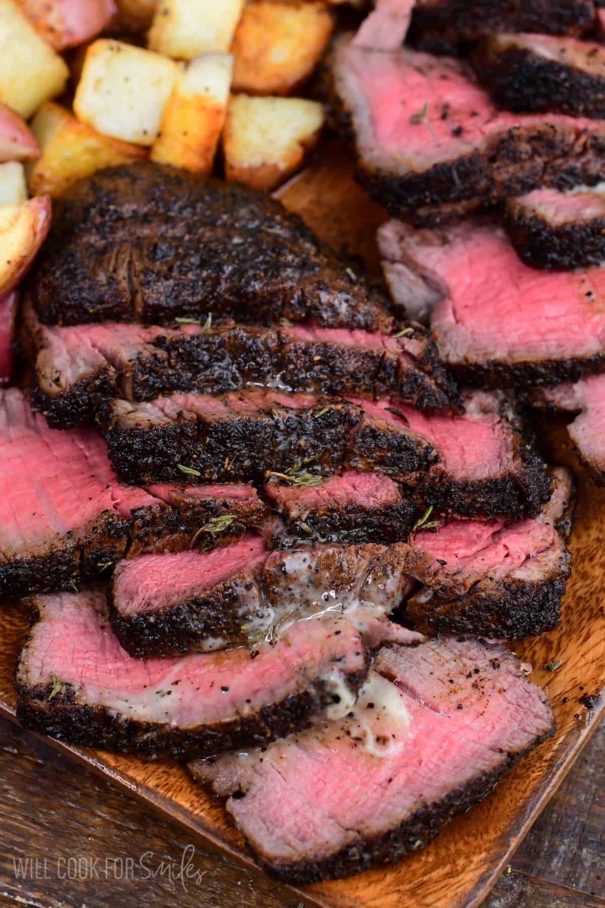 Sliced coffee-rubbed filet mignon arranged on a wooden serving platter, featuring a tender medium-rare center and a rich, dark crust. Melted herb butter and savory juices coat the steak slices, while crispy roasted potatoes add a hearty side dish in the background.