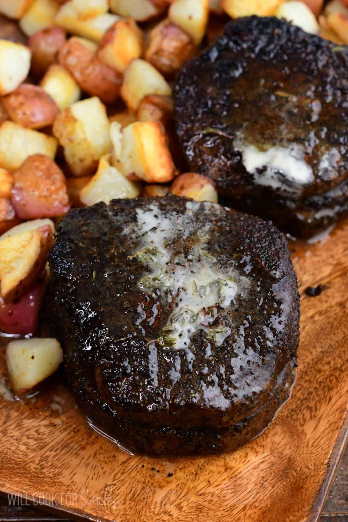 Two coffee-rubbed filet mignon steaks topped with melted herb butter, served alongside roasted red potatoes on a wooden platter.