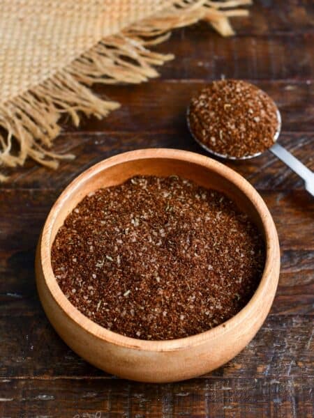 Coffee steak rub seasoning blend in a wooden bowl with a measuring spoon filled with the spice mixture on a rustic wooden table.
