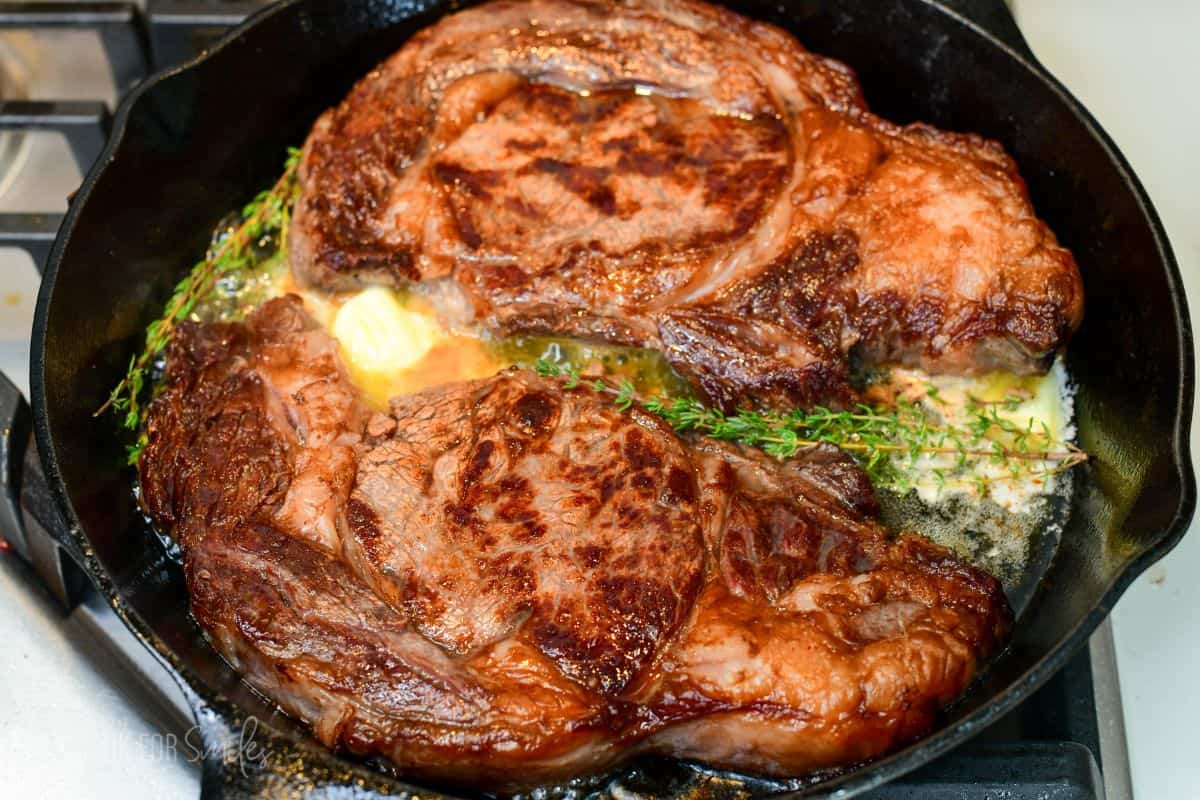 Two ribeye steaks searing in a cast-iron skillet with butter and fresh thyme, developing a rich golden-brown crust.