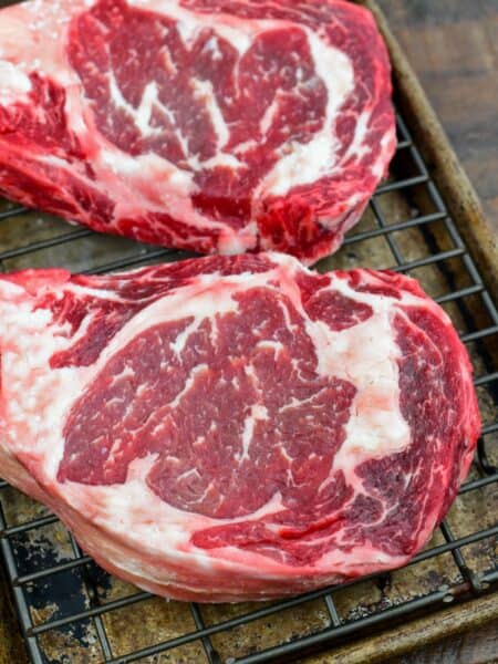 Two ribeye steaks after dry brining, resting on a wire rack with a deep red color and marbling before cooking.