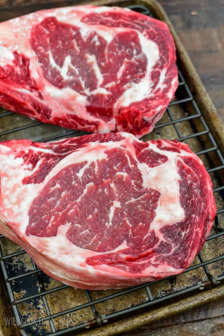 Two ribeye steaks after dry brining, resting on a wire rack with a deep red color and marbling before cooking.