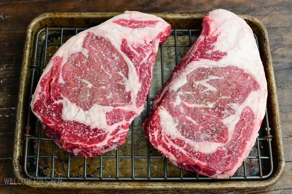 Two dry brined ribeye steaks seasoned with coarse salt, resting on a wire rack over a baking sheet before cooking.