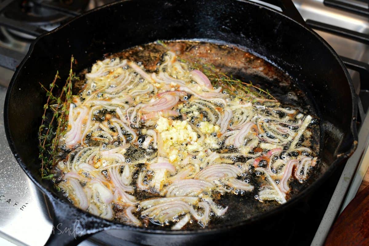 Shallots, garlic, and fresh thyme simmering in butter and steak drippings in a cast-iron skillet to make pan sauce.