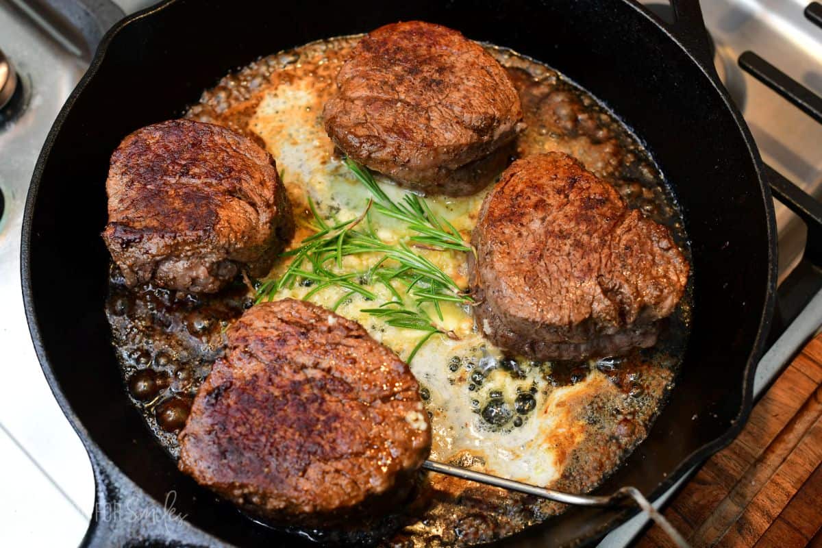 Four filet mignon steaks finish cooking in a cast-iron skillet, surrounded by sizzling butter, garlic, and fresh rosemary.
