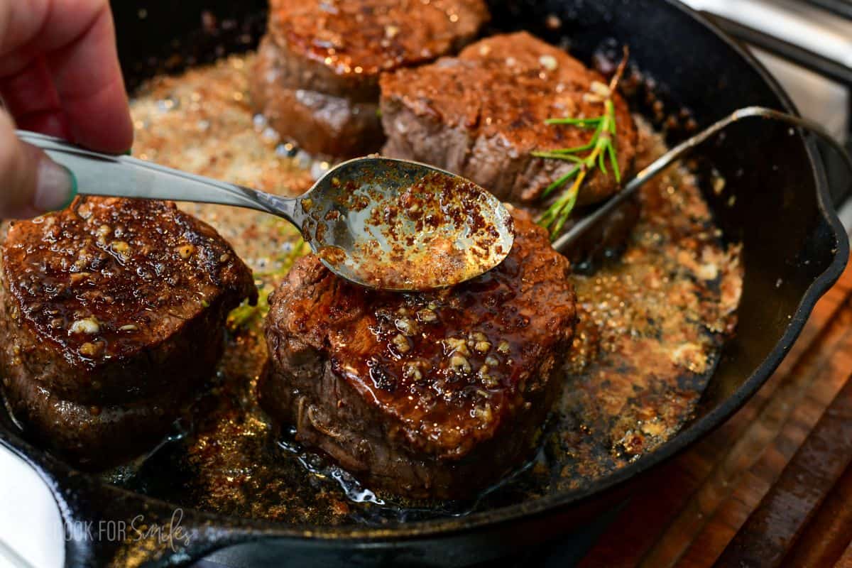 A spoon drizzles sizzling garlic-herb butter over filet mignon steaks in a cast-iron skillet, basting the beef as it finishes cooking. The steaks have a beautifully browned crust, while rosemary, garlic, and rich pan juices add flavor and help keep the meat tender and juicy.