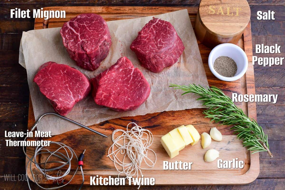 Labeled ingredients for pan-seared filet mignon arranged on a wooden cutting board, including raw filet mignon steaks, butter, garlic cloves, fresh rosemary, salt, black pepper, kitchen twine, and a leave-in meat thermometer.