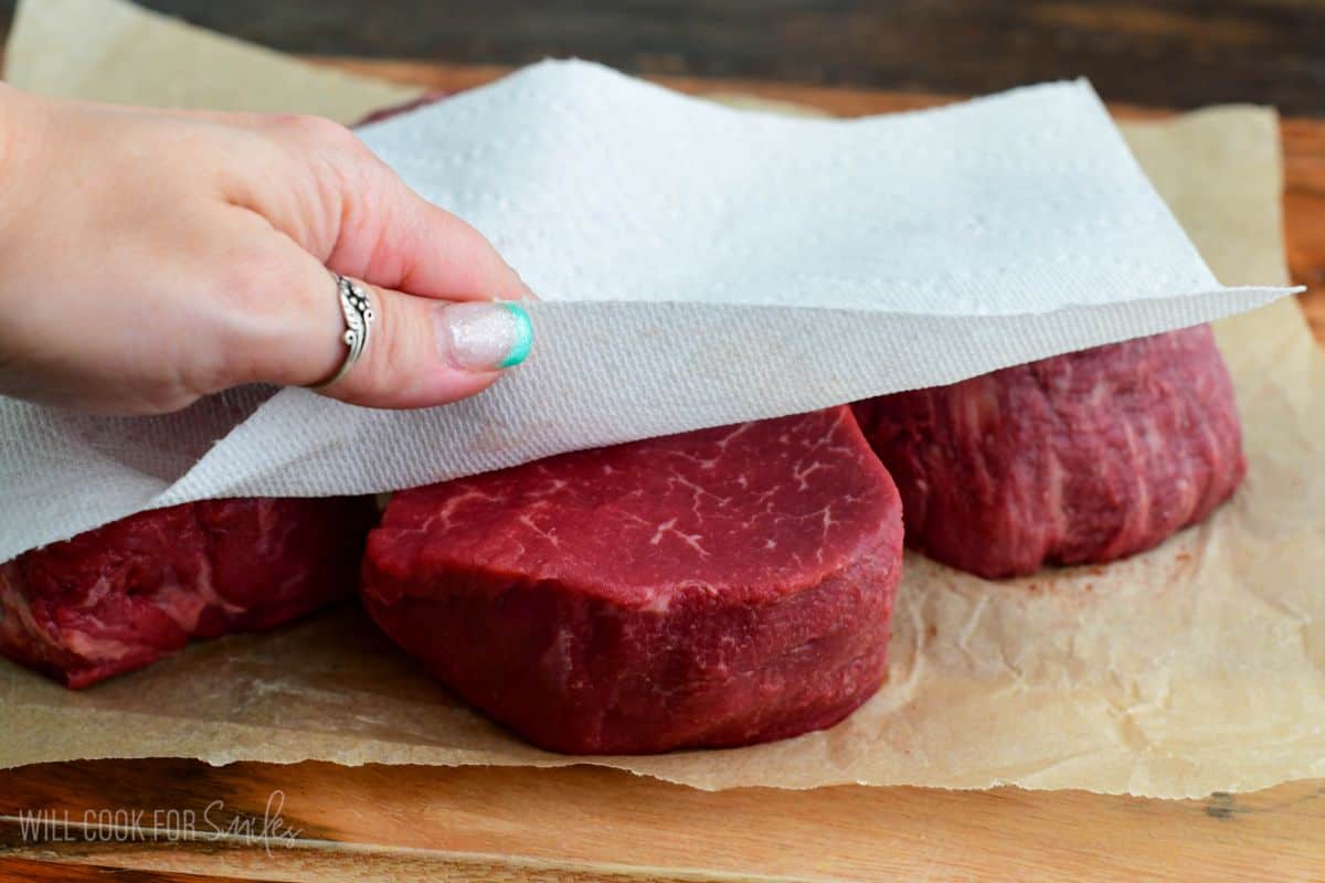 Patting dry raw filet mignon steaks with a paper towel on parchment paper, helping remove excess moisture before seasoning and cooking.