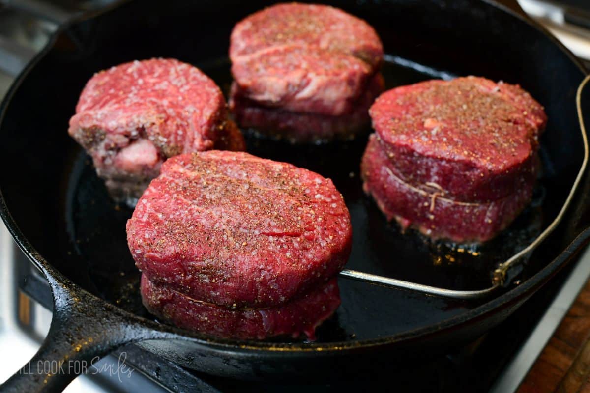 Four seasoned filet mignon steaks sear in a cast-iron skillet, developing a flavorful crust. One steak has a thermometer probe inserted to monitor the internal temperature.
