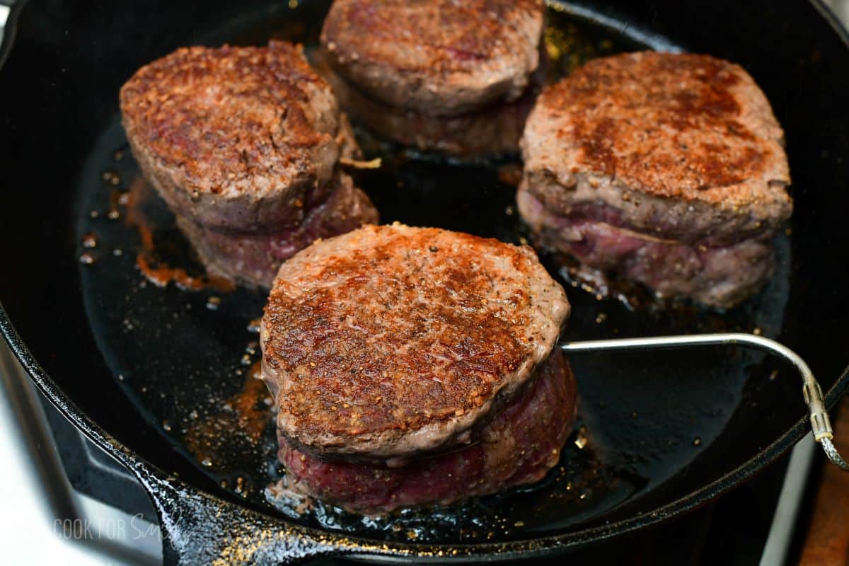 Four filet mignon steaks sear in a cast-iron skillet, showing a rich golden-brown crust forming on the surface. A temperature probe is inserted into one steak to track doneness.