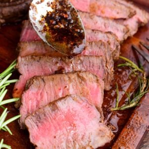Sliced filet mignon arranged on a wooden cutting board, showing a juicy medium-rare center. A spoon drizzles garlic-herb butter and pan juices over the steak.