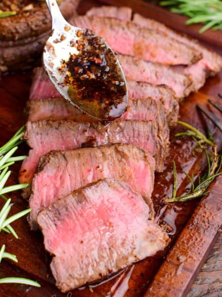 Sliced filet mignon arranged on a wooden cutting board, showing a juicy medium-rare center. A spoon drizzles garlic-herb butter and pan juices over the steak.