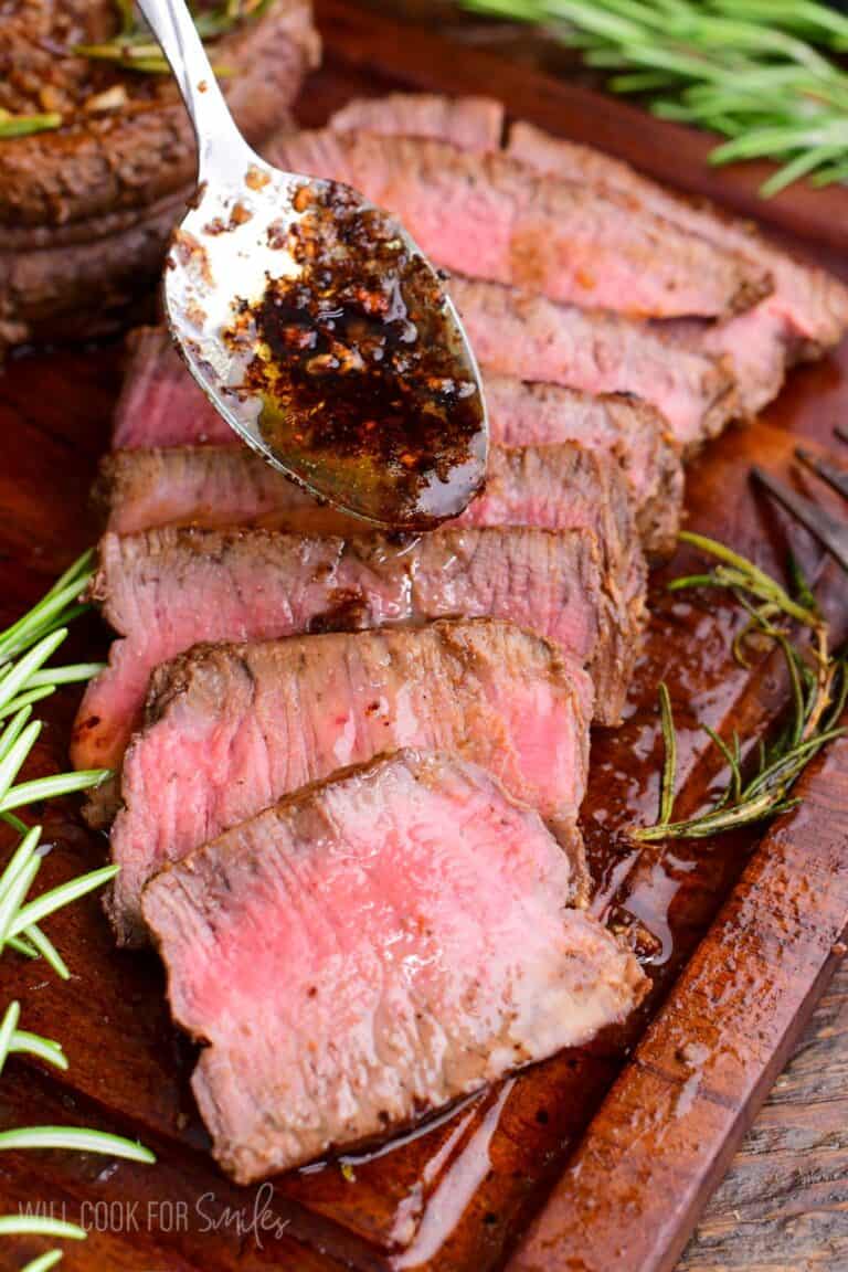 Sliced filet mignon arranged on a wooden cutting board, showing a juicy medium-rare center. A spoon drizzles garlic-herb butter and pan juices over the steak.