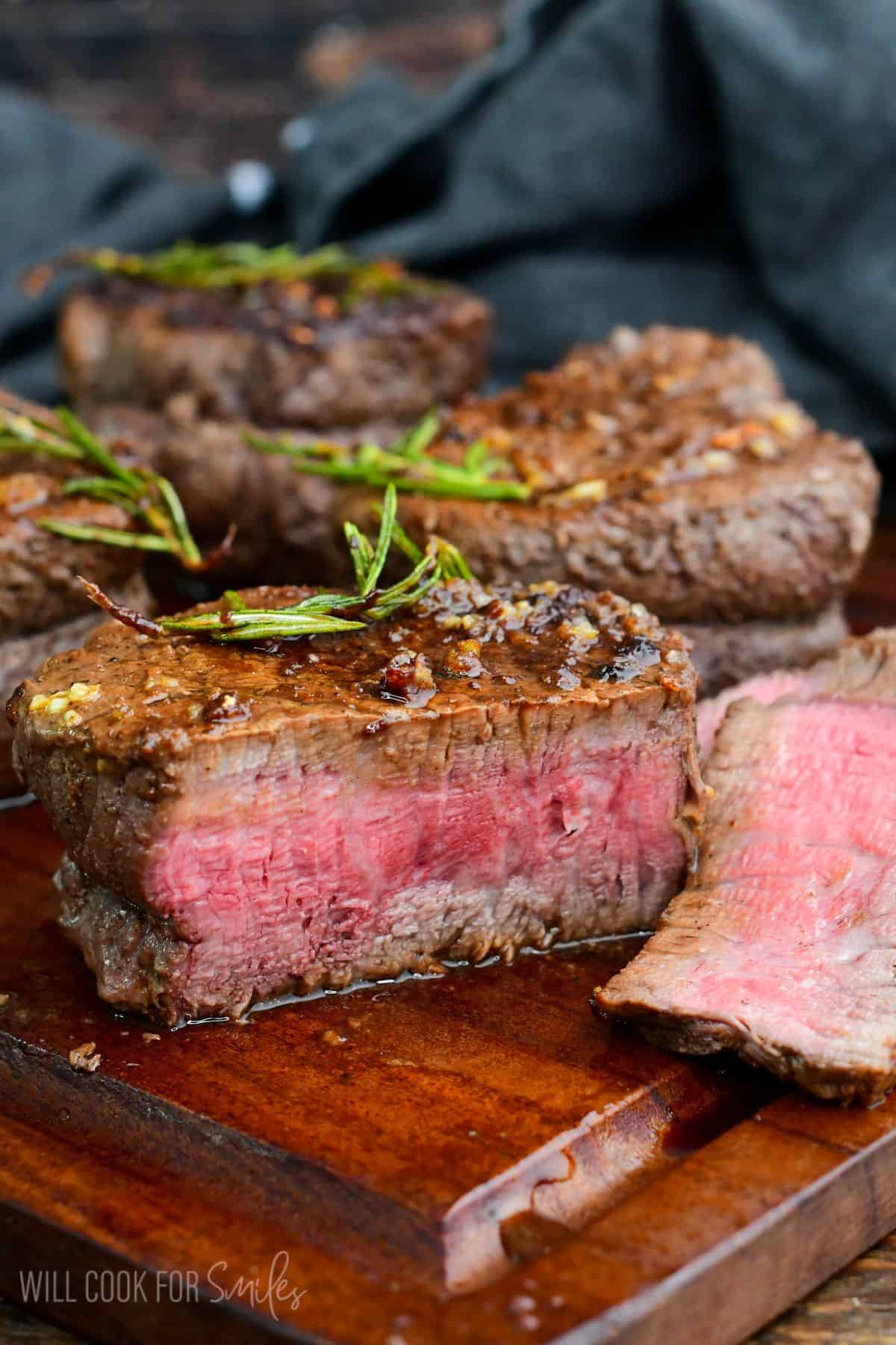 filet mignon sliced open to reveal a warm pink medium-rare to medium center, resting on a wooden cutting board. The steak is topped with rosemary and garlic butter, with savory pan juices pooling around it.