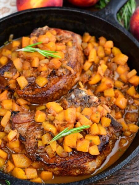Peach pork chops simmered in a cast-iron skillet with diced peaches and savory peach sauce, garnished with rosemary.