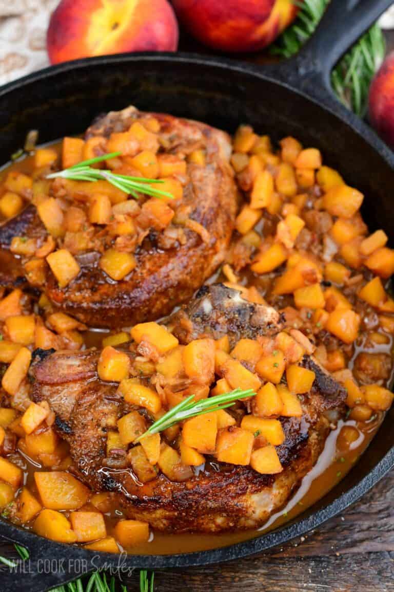 Peach pork chops simmered in a cast-iron skillet with diced peaches and savory peach sauce, garnished with rosemary.