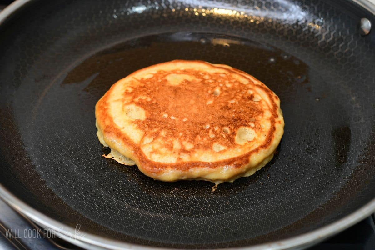 Golden brown banana pancake cooking in a nonstick skillet after flipping, with an evenly browned surface.