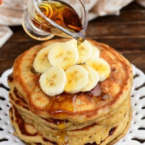 Maple syrup being poured over a stack of fluffy banana pancakes topped with fresh banana slices on a serving plate.