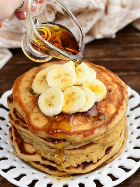 Maple syrup being poured over a stack of fluffy banana pancakes topped with fresh banana slices on a serving plate.
