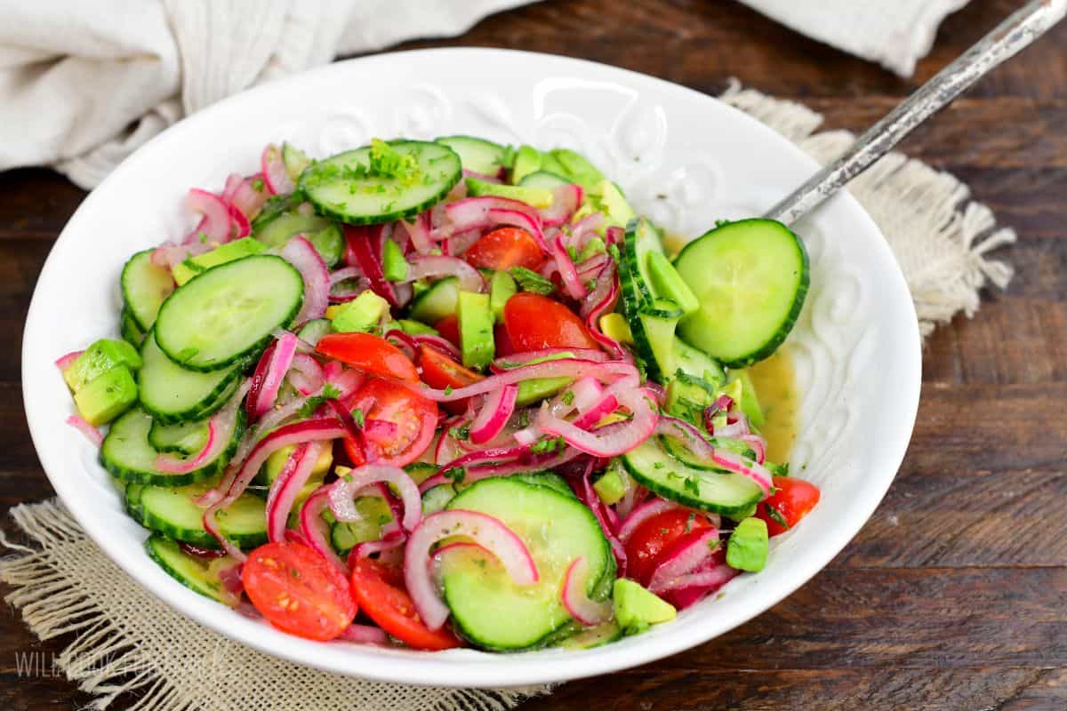 Cucumber tomato onion salad with avocado, grape tomatoes, fresh herbs, and vinaigrette in a white bowl.