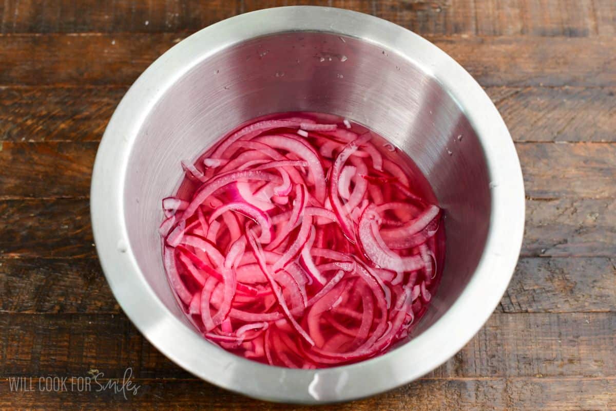 Thinly sliced red onions soaking in pickling liquid in a stainless steel bowl.