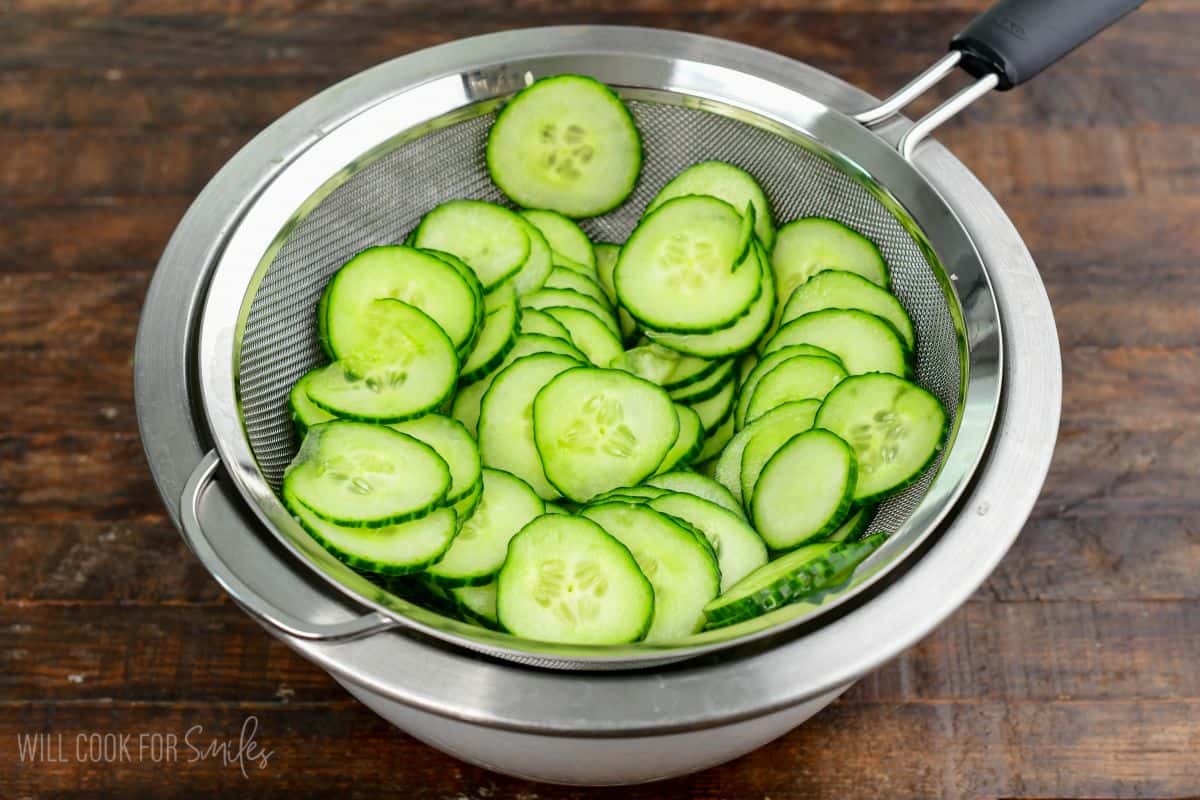Thinly sliced cucumbers draining in a mesh strainer over a stainless steel bowl.