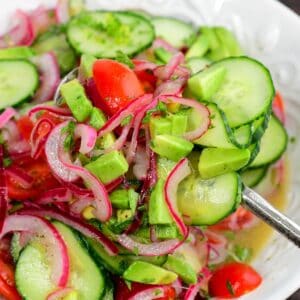 Cucumber tomato onion salad with avocado and fresh herbs being scooped from a bowl with a serving spoon.