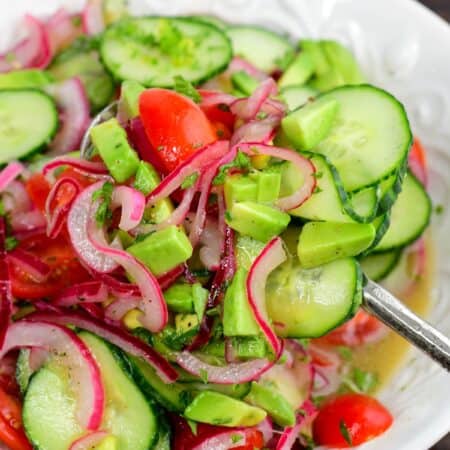 Cucumber tomato onion salad with avocado and fresh herbs being scooped from a bowl with a serving spoon.